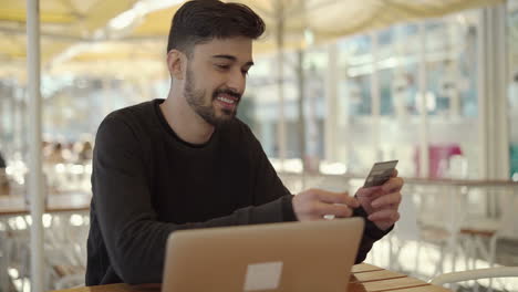 Smiling-man-holding-credit-card-and-using-laptop-in-cafe
